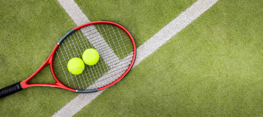 A red tennis racquet and two bright yellow tennis balls on a green court, illustrating spring pre-season gear preparation.